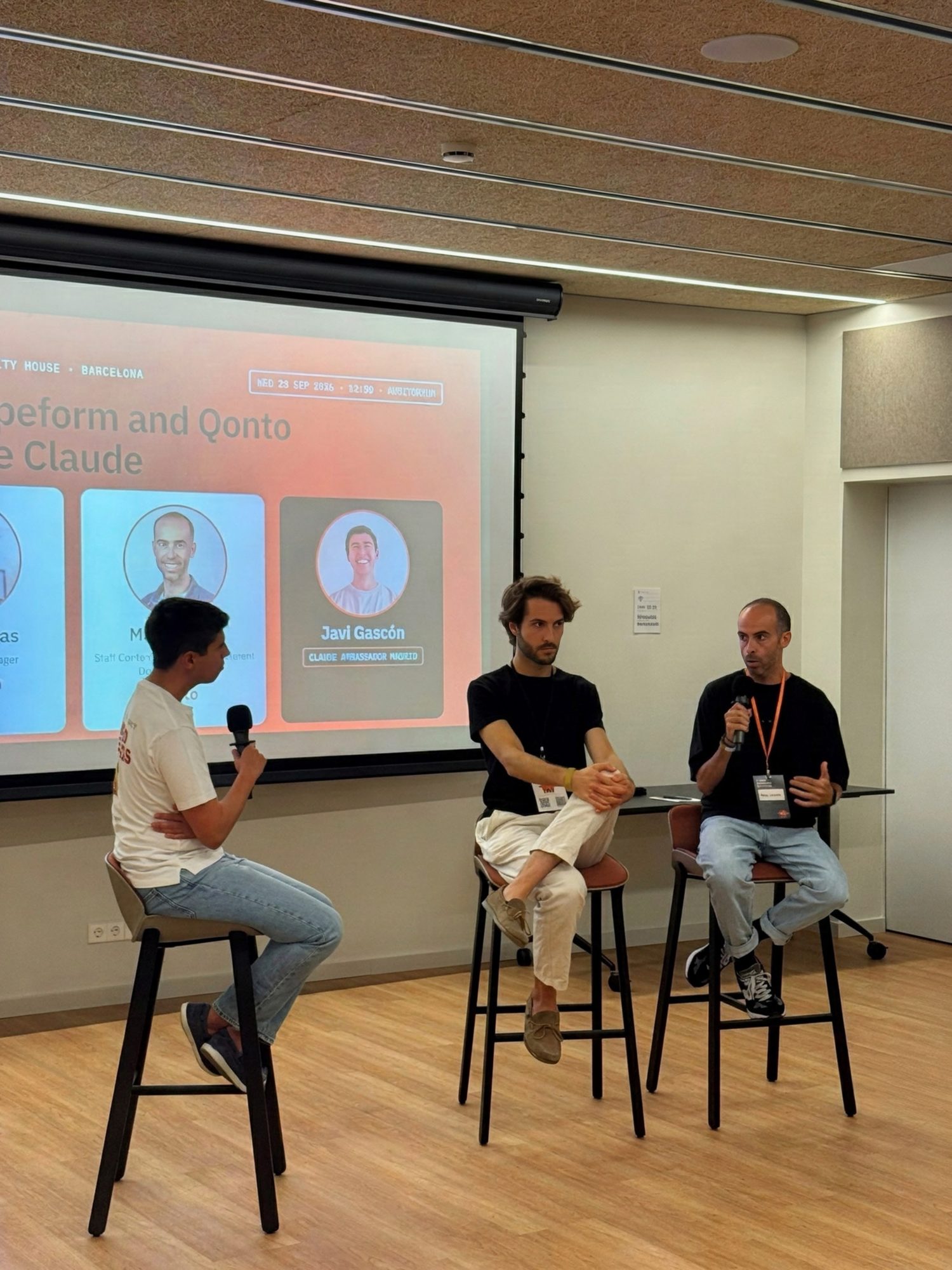 Three speakers on stools in front of a projected slide titled 'How Typeform and Qonto leverage Claude', two of them holding microphones, at Claude Community House Barcelona